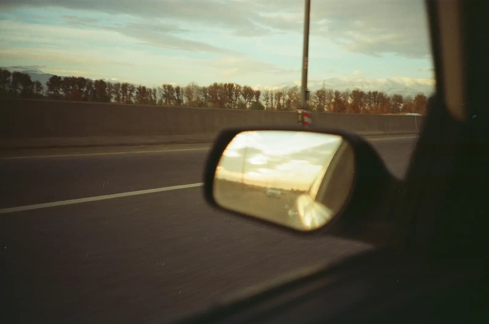 View from a moving car: the side mirror reflects a golden sunset over the road behind
